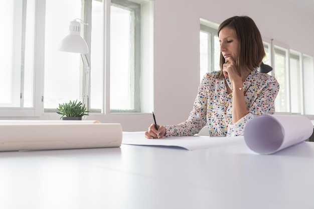 Jeune femme assise dans un bureau travaillant sur un projet avec des dessins ou des plans enroulés sur son bureau.