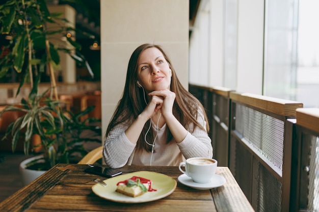 Jeune femme assise dans un café à table avec une tasse de cappuccino, gâteau, se détendre au restaurant pendant le temps libre. Une jeune femme écoute de la musique dans des écouteurs sur un téléphone portable, se repose au café. Concept de mode de vie.