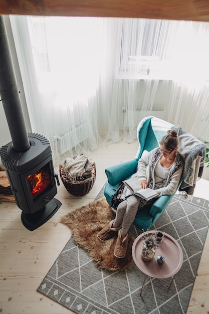 Jeune femme assise dans un fauteuil confortable avec une couverture chaude en lisant un livre