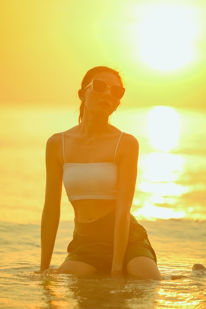 Jeune femme assise dans le sable à la mer au coucher du soleilFemme profitant de la plage relaxante joyeuse en été avec le coucher du soleil