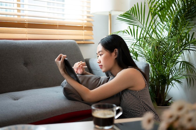 Jeune femme assise dans le salon et naviguant sur Internet sans fil ou faisant du shopping sur une tablette numérique