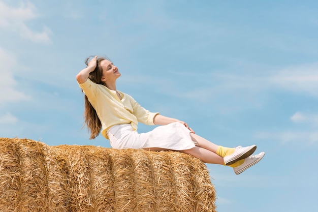 Jeune femme assise sur de grandes piles de paille Modèle féminin posant dans le champ après la récolte par la botte de foin