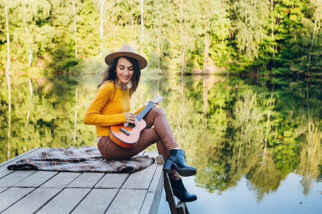 Jeune femme assise avec une guitare sur un pont sur un lac avec un paysage d'automne. Tonifiant.