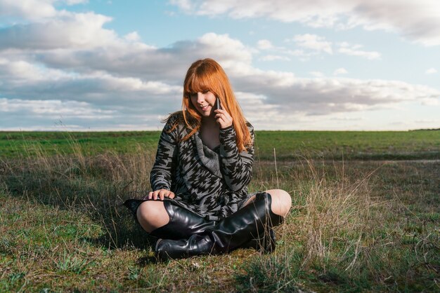 Jeune femme assise sur l'herbe verte du champ parlant au téléphone sous un ciel nuageux