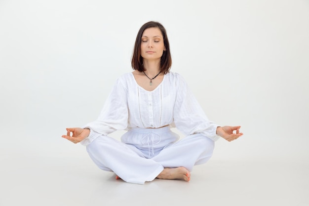 Jeune femme assise avec les jambes croisées sur le sol faisant de l'exercice Sukhasana avec les yeux fermés sur fond blanc Yoga