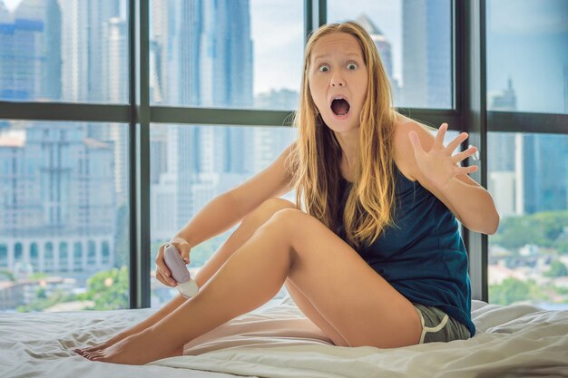 Jeune femme assise sur le lit à la maison et faisant de l'épilation avec un épilateur sur les jambes et souffre. Sur le fond d'une fenêtre donnant sur la grande ville, des gratte-ciel