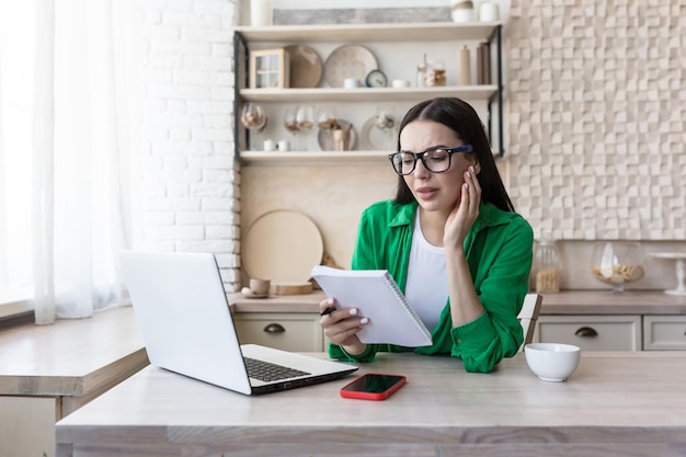 Une jeune femme assise à la maison dans la cuisine avec un ordinateur portable note le budget familial dans un carnet