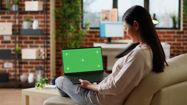 Jeune Femme Assise à La Maison Avec Un Ordinateur Portable Ayant Un Affichage à écran Vert Chroma Key. Personne Adulte Assise Sur Un Canapé Avec Un Ordinateur Portable Sur Les Genoux Ayant Une Maquette De Fond De Modèle Isolé. Prise De Vue Au Trépied