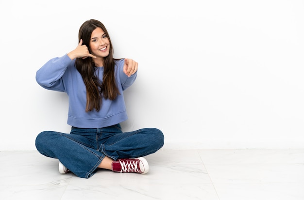 Jeune femme assise par terre faisant un geste de téléphone et pointant vers l'avant