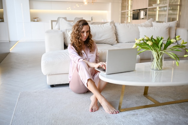 Photo jeune femme assise près d'un canapé blanc dans un intérieur de salon de style scandinave travaillant sur ordinateur portable.