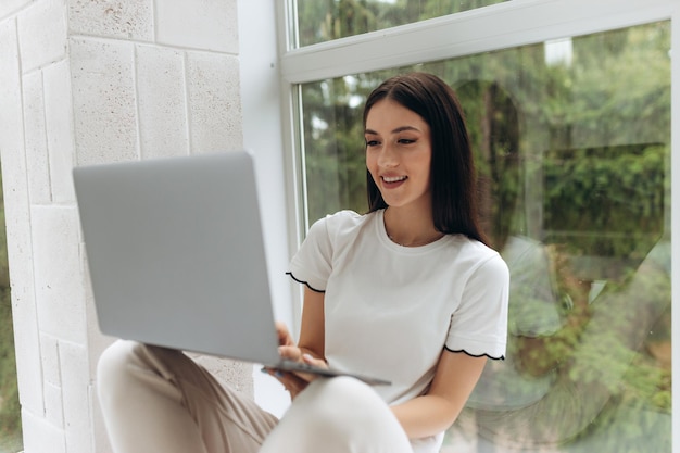 Jeune femme assise sur le rebord de la fenêtre et travaillant en ligne à l'aide de son ordinateur portable dans la chambre