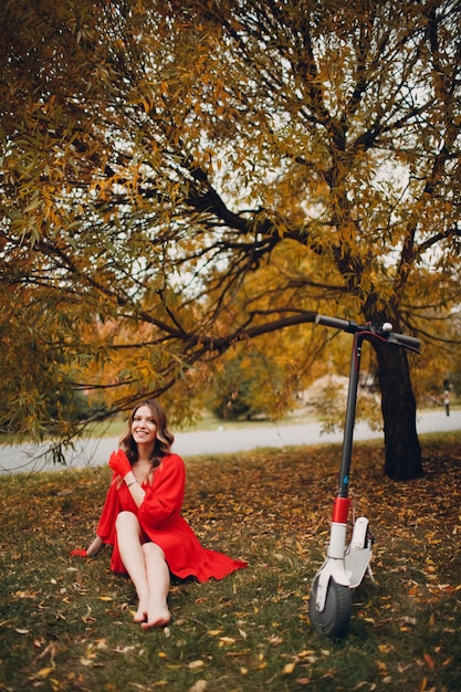 Jeune femme assise avec scooter électrique en robe rouge au parc de la ville d'automne