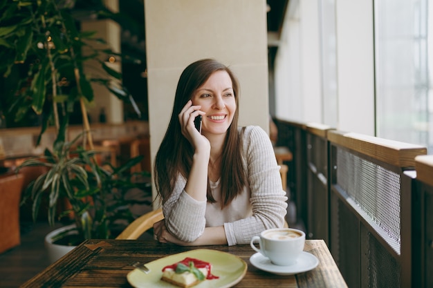 Jeune femme assise seule dans un café à table avec une tasse de cappuccino, gâteau, détente au restaurant pendant le temps libre. Jeune femme parlant au téléphone mobile, se reposant au café. Concept de mode de vie.