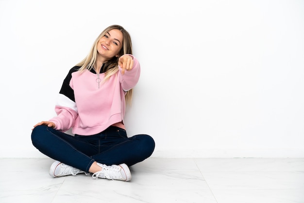 Jeune femme assise sur le sol à l'intérieur pointant vers l'avant avec une expression heureuse