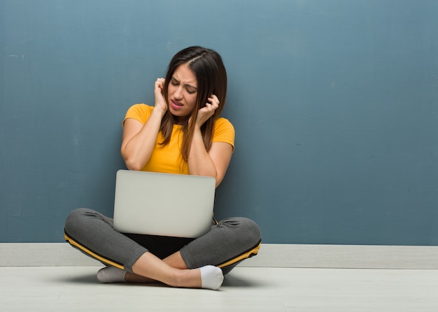 Jeune femme assise sur le sol avec un ordinateur portable couvrant les oreilles avec les mains