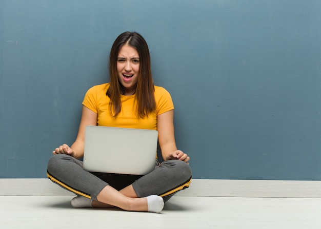 Jeune femme assise sur le sol avec un ordinateur portable hurlant très en colère et agressive