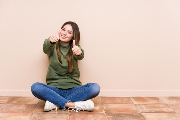 Jeune femme assise sur le sol avec les pouces vers le haut, applaudit à propos de quelque chose, de soutien et de respect du concept.