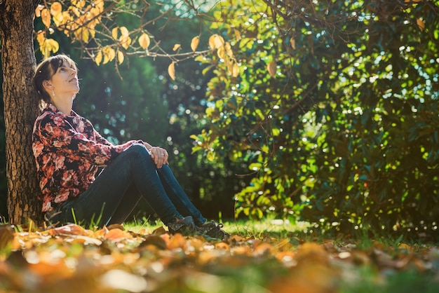 Jeune femme assise sous un arbre d'automne s'appuyant sur le tronc d'arbre profitant et se relaxant.