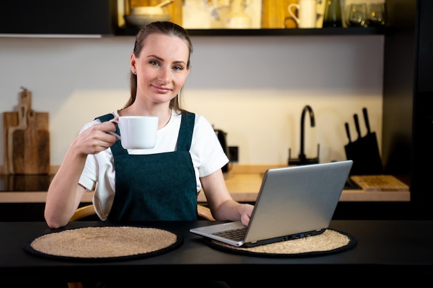 Jeune femme assise à la table de la cuisine travaillant sur un ordinateur portable en buvant du café