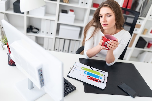 Une jeune femme assise à une table dans le bureau et tenant une tasse rouge.