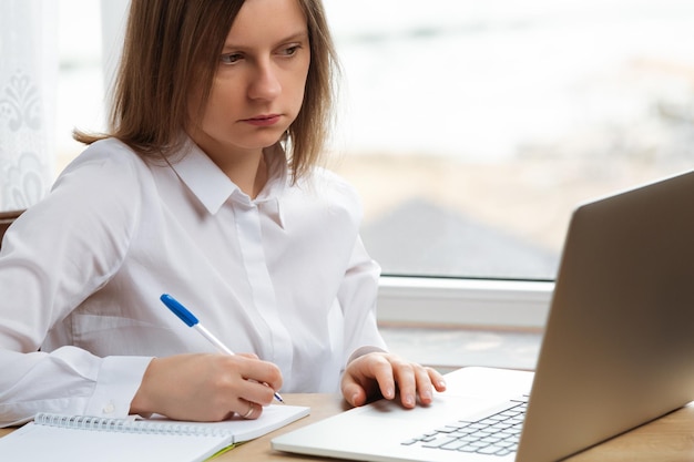 Jeune femme assise à une table de travail avec ordinateur portable et bloc-notes pour travailler à domicile