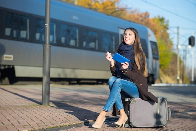 Jeune femme assise sur une valise à l'aide d'une tablette à la gare