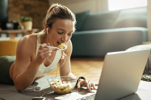 Jeune femme athlétique allongée sur le sol et mangeant des aliments sains tout en surfant sur le net sur un ordinateur portable.