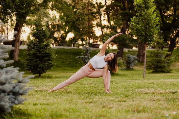 Jeune femme athlétique pratique le yoga dans un parc verdoyant sur l'herbe le matin d'été Tenez-vous debout dans la pose d'angle latéral étendu sur fond de forêt de vêtements de sport Concept de mode de vie sain et d'harmonie avec le corps