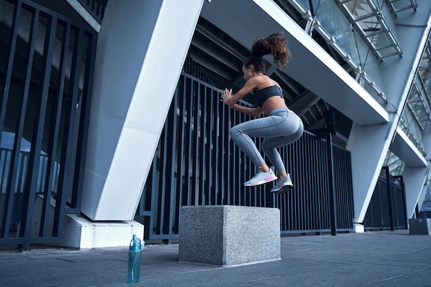 Jeune femme athlétique sautant sur un cube tout en s'entraînant au stade de la ville