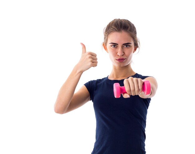 Jeune femme athlétique en T-shirt bleu tenant une haltère rose et montrant le pouce vers le haut sur fond blanc en studio