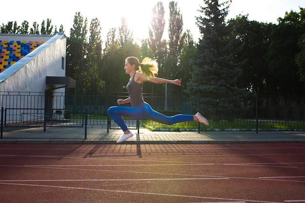 Jeune femme athlétique en t-shirt gris et leggings bleus sautant sur la piste du stade. Espace pour le texte