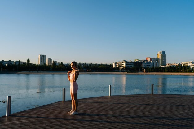 Jeune femme athlétique en vêtements de sport faisant de l'exercice et s'étirant musculairement pour s'échauffer avant de courir ou de s'entraîner au bord de l'eau dans le parc de la ville