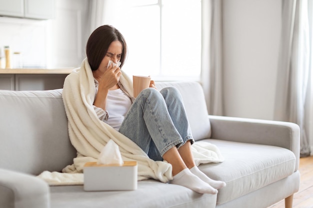 Une jeune femme atteinte de rhume et de grippe se mouche le nez et boit du thé.