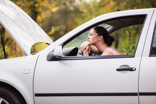 Une jeune femme attend de l'aide près de sa voiture, qui est tombée en panne sur le bord de la route