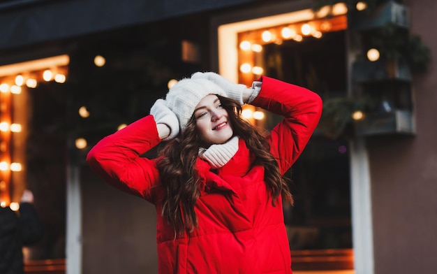 Une jeune femme attirante dans une veste d'hiver rouge et des mitaines pose dans le contexte de la ville du soir