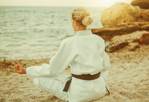 Jeune femme attirante de karaté dans le kimono blanc avec une méditation de ceinture noire sur la plage sauvage avec des pierres