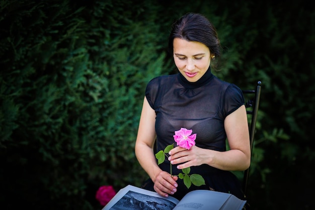 Jeune femme attirante s'asseyant dans un jardin avec un grand livre et une fleur rose dans des ses mains