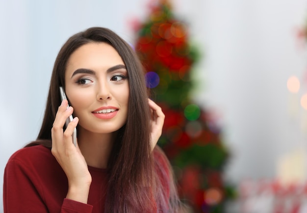 Jeune femme attirante avec le téléphone portable sur le fond brouillé de Noël, à l'intérieur