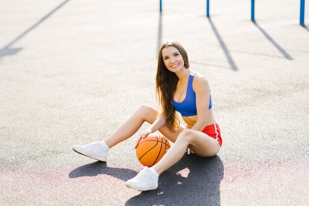 Photo une jeune femme attrayante et athlétique posant en plein air assise sur un terrain de basket-ball tenant une personne de basket-bal