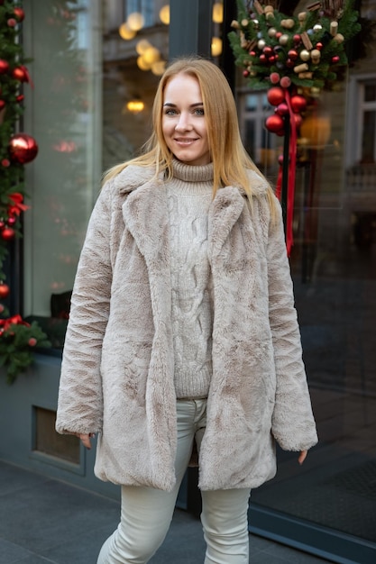 Une jeune femme attrayante dans un manteau de fourrure près du magasin Décorations de Noël à l'arrière-plan Marche de la saison des fêtes dans la ville