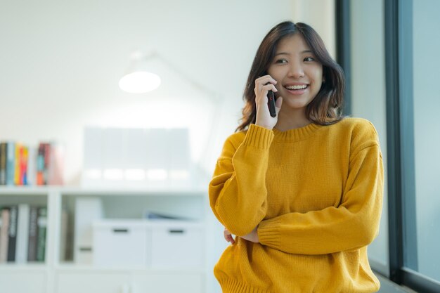 Photo une jeune femme attrayante utilisant un smartphone se tient près de la fenêtre dans une pose décontractée se sentant contemplative dans son bureau à la maison