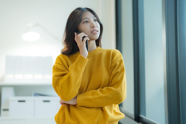 Une jeune femme attrayante utilisant un smartphone se tient près de la fenêtre dans une pose décontractée se sentant contemplative dans son bureau à la maison