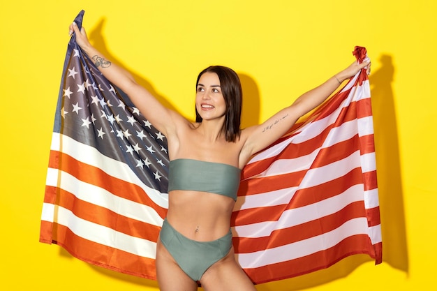Une jeune femme au bronzage doré et au sourire radieux pose dans un maillot de bain chic avec un drapeau américain sur fond jaune vif L'image capture l'essence de vacances d'été glamour