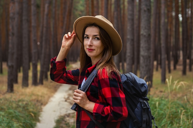 Jeune femme au chapeau, chemise rouge et sac à dos dans la forêt. Randonnée en forêt.