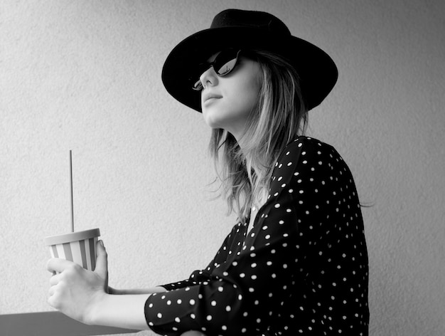 Jeune femme au chapeau et lunettes de soleil avec un verre. Image en style noir et blanc