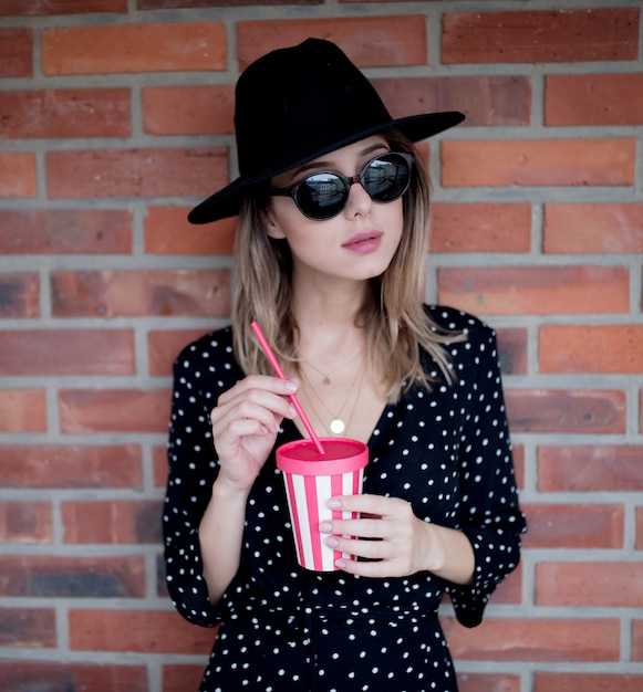 Jeune femme au chapeau et lunettes de soleil avec un verre