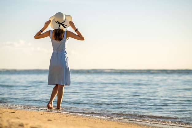 Jeune femme au chapeau de paille et une robe debout seule sur la plage de sable vide au bord de la mer