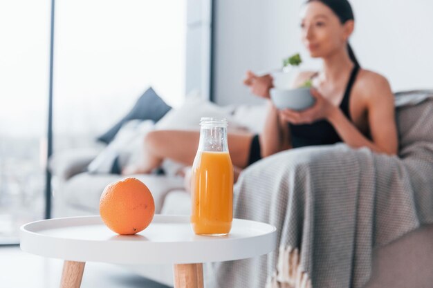 Une jeune femme au corps mince en vêtements de sport est assise sur un canapé et mange des aliments sains à l'intérieur à la maison