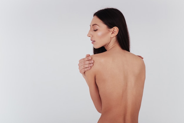 Une jeune femme au corps parfait et à la peau lisse se tient dos au mur blanc.