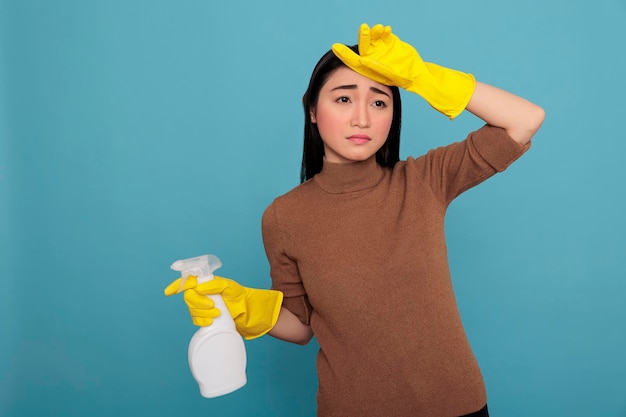 Jeune femme au foyer asiatique stressée par le travail quotidien tenant un détergent en aérosol dans un gant en caoutchouc jaune Concept de maison de nettoyage, femme de ménage frustrée par le surmenage avec un état d'esprit négatif